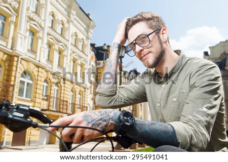Handsome self-confident hipster guy is touching his hair flirtingly. He is sitting on bicycle. He has tattoo and glasses