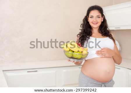 Beautiful pregnant woman prefers healthy eating. She is showing a bowl of fruits and smiling with joy. The lady is touching her abdomen with love. Copy space in left side