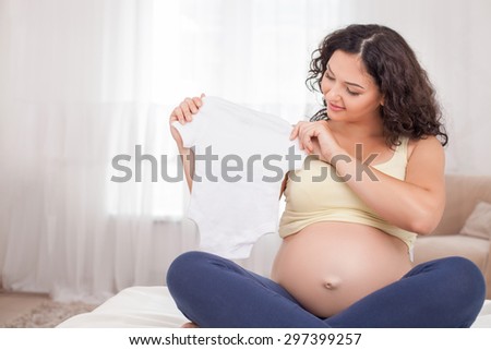 Beautiful pregnant woman is holding a small T-shirt in her hands. She is looking at it and smiling. The lady is sitting in lotus position on bed. Copy space in left side