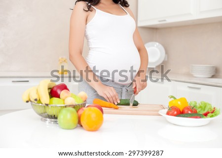 Close up of body of pregnant woman standing in the kitchen and cutting cucumber. There are a lot of fruits and vegetable on the table