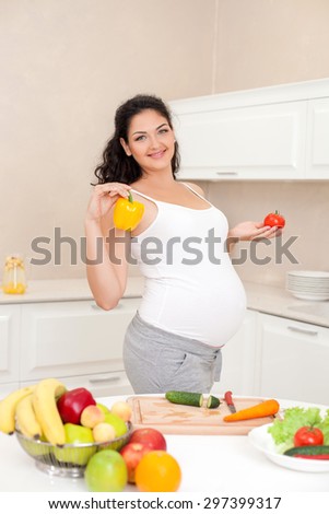 Cute pregnant woman is standing and holding vegetables. She is smiling with enjoyment. There are many healthy fruits and vegetables on the table