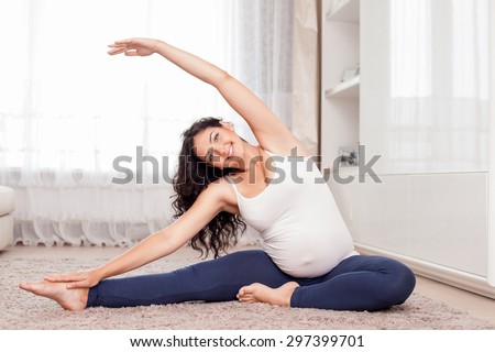 Pretty expectant mother is exercising in her room. She is sitting on floor and stretching her arms aside. The lady is smiling and looking at the camera with joy