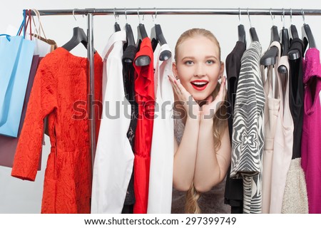 Attractive blond girl is standing between dresses hanging on rack. She is raising her hand to her cheek and smiling happily