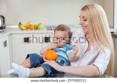 Cute mom is proposing orange to her small son. He is looking at it with appetite. The woman is sitting at the table and holding the boy on her knees. She is smiling