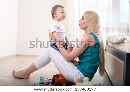 Cheerful mother is teaching her male toddler to talk. She is holding him and sitting on flooring. The mom is looking at her child with love