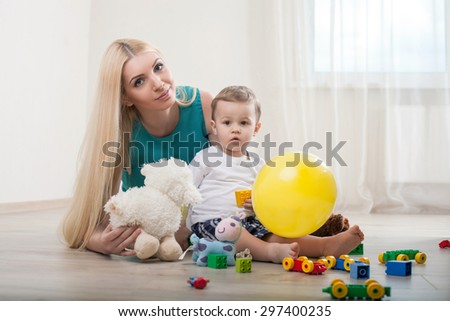 Cute mom and her child are playing with toys on floor. The woman is looking forward and smiling. Her son is staring at something thoughtfully