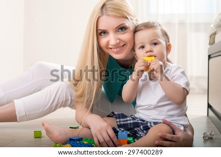 Pretty mother is sitting near her son and embracing him. They are looking at the camera with interest. The toddler is trying to bite the toy. His mamma is smiling