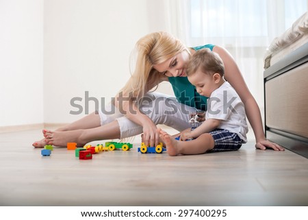 Cheerful mom is playing with her kid in bedroom. The boy is looking at toys with interest. His mamma is smiling happily