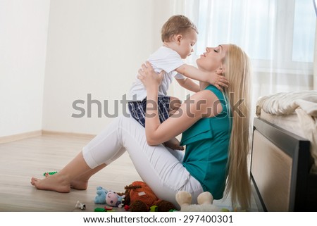 Beautiful mom is hugging her small son with love. They are looking at each other gently. The parent is holding her kid and sitting on floor