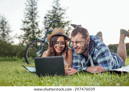 Pretty young loving couple is lying on cover in nature. The are watching film from notebook with interest. The lovers are drinking wine and smiling