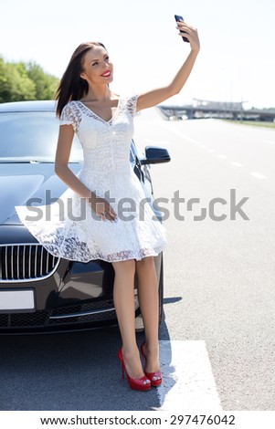 Beautiful girl is leaning on her car and making selfie. She is smiling and looking at the telephone with happiness. Copy space in right side
