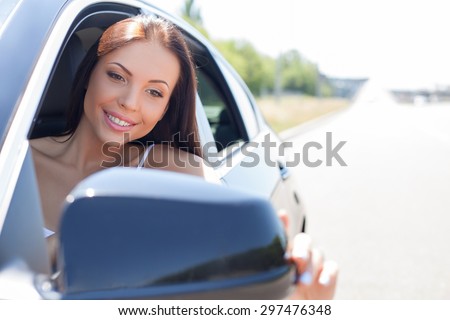 Attractive woman is looking at side mirror of her car and adjusting it with her hand. The lady is smiling with satisfaction. She is sitting at seat of driver. Copy space in right side