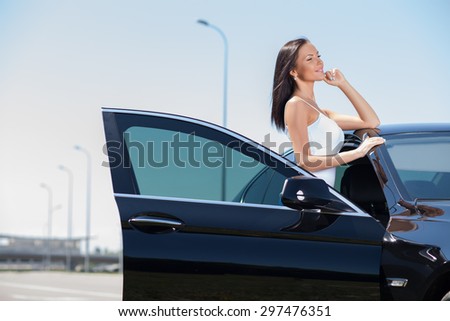 Pretty girl is standing near her car. She is leaning on the roof and is looking aside dreamingly. She is smiling with anticipation. Copy space in left side