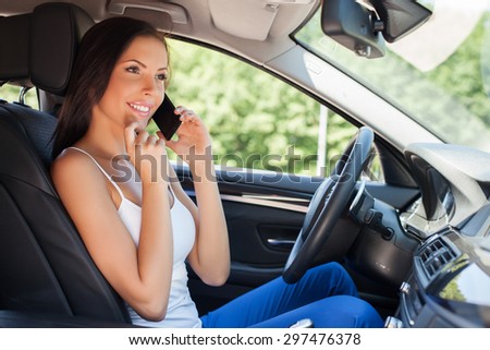 Attractive girl is sitting at steering wheel in her new car. She is talking on the telephone. The lady is touching her chin with interest and smiling