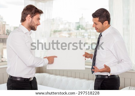 Attractive healthy businessmen are holding a white board and looking at it with concentration. They are standing and discussing some concepts. The man is pointing his finger at it and smiling