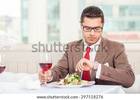 Handsome man in suit is sitting at the table and looking at his watch attentively. He is waiting for his business partner. The man is drinking wine and eating a salad. Copy space in left side