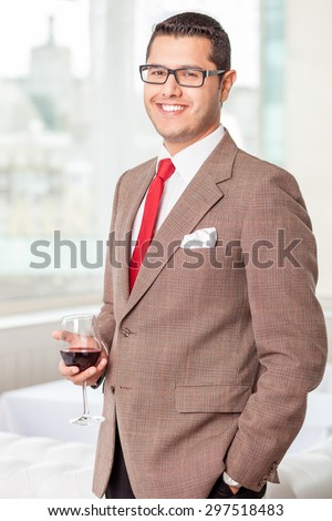 Cheerful businessman is standing and drinking red wine. He is smiling and looking at the camera with happiness