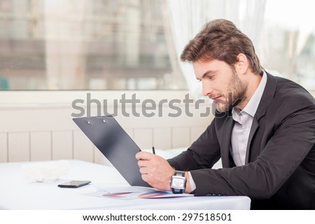 Cheerful man is reading demands of a new project with concentration. He is looking at the folder of documents seriously. The man is sitting at the table. Copy space in left side
