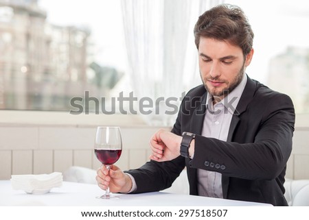 Handsome man is sitting in restaurant and looking at his watch seriously. He has an appointment with his business partner. He is drinking red wine. Copy space in left side