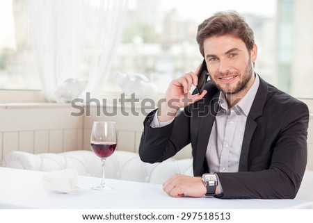 Cheerful businessman is talking on mobile phone in the restaurant. He is arranging a meeting with his client. He is smiling and looking at the camera confidently. Copy space in left side