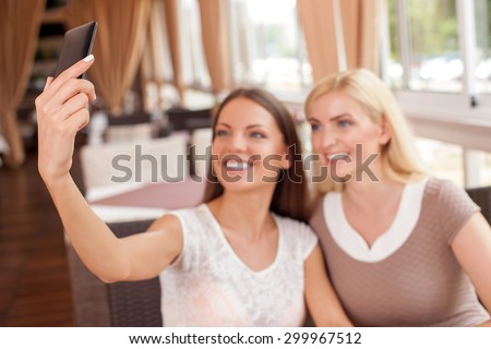 Attractive girls are sitting in cafeteria and photographing themselves. The women are smiling with joy. Focus on female hand with a mobile phone