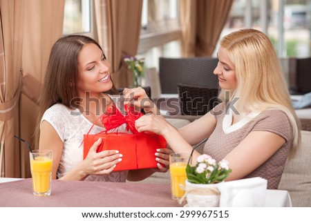 Cheerful women are sitting in cafe. The brunette girl is holding a gift and looking at her friend mysteriously. The blond lady is opening the gift with interest. They are smiling with joy