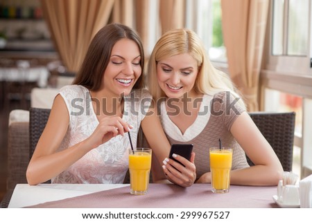 Cheerful woman are sitting at the table in cafe. They are drinking juice. One lady is holding a mobile phone. The friends are looking at it with interest and smiling