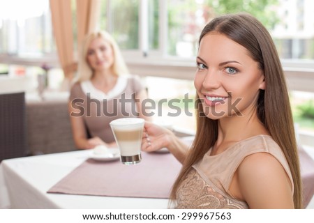 Attractive two women are sitting at the table in cafe. They are drinking latte with enjoyment. Focus on brunette girl. She is holding the cup and smiling