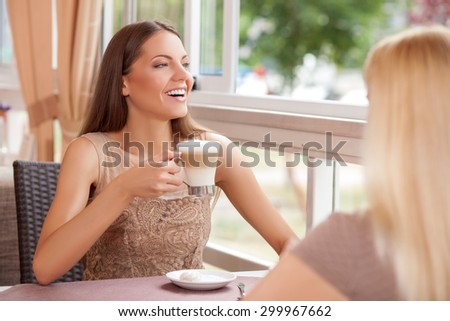 Pretty women are talking and drinking latte in restaurant. The girls are sitting at the table opposite each other. They are looking through the window and laughing