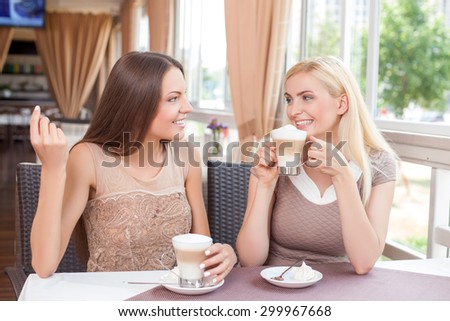 Beautiful women are drinking latte together in cafe. The friends are talking and smiling. One woman is gesturing with joy