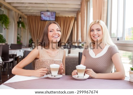 Pretty women are sitting at the table and drinking latte in cafe. They are spending time with joy. The women are smiling and looking at the camera with enjoyment