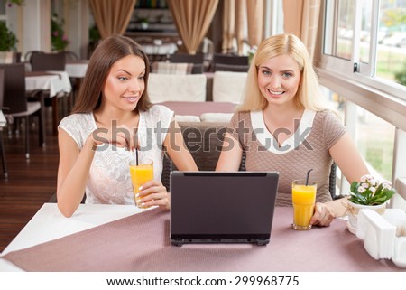 Beautiful girls are resting in cafe. They are drinking juice and smiling. The women are using an internet and looking at the notebook with curiosity and surprise