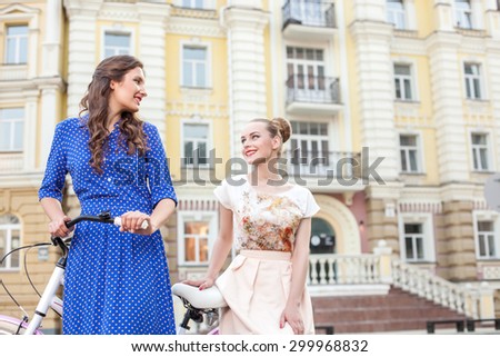 Pretty women are cycling in town with joy. They are looking at each other and smiling. The brunette lady is riding a bicycle and her friend is sitting on it behind her. Copy space in right side