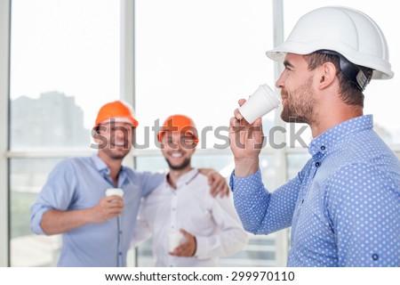 Cheerful construction team are making coffee break. The architect is drinking coffee with pleasure and smiling. Two workers are looking at him and laughing. They are embracing friendly