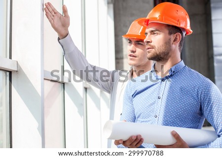 Cheerful young foreman is showing to the architect the problems of construction. He is pointing his arm sideways. The men are looking aside with seriousness