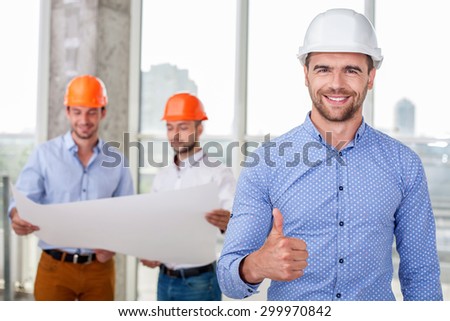 Attractive architect is giving thumb up. The man is smiling and looking at the camera with happiness. His plan was approved. Two builders are looking at sketches of building on background