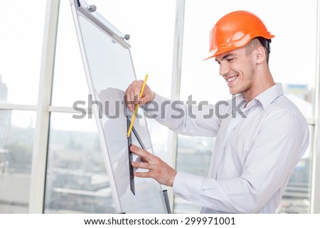 Handsome builder is drawing sketches of construction on vertical board. He is smiling and looking at his work with enjoyment