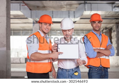 Pretty construction team is ready to sign a contract. The old architect is showing a folder of document. The young worker is pointing his finger at it. The men are smiling