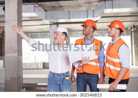 Senior architect is expressing his ideas concerning building. He is pointing his arm sideways and smiling. The man is holding a blueprint. The builders are looking aside with interest and joy