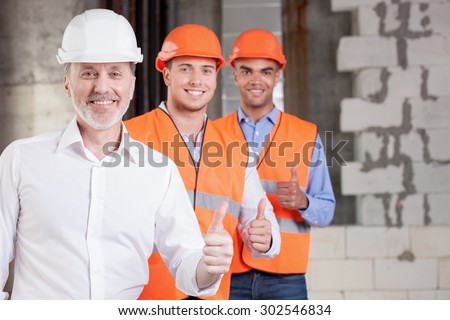 Skilled old architect and young workers are giving thumbs up. Their project was approved. The men are smiling and looking at the camera with happiness. Copy space in right side
