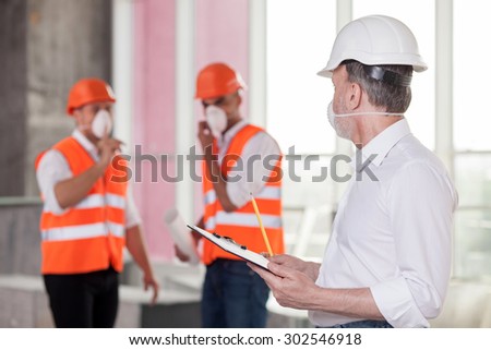 Experienced old architect is discussing with builders the concepts of building. He is writing down their ideas and looking at them with interest. The men are wearing helmets and respirators