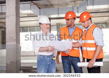 Skilled old architect is explaining to workers his ideas about a project with aspirations. He is smiling. The men are looking at blueprint with surprise. Copy space in left side