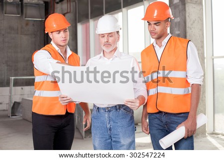 Experienced old architect is holding blueprint. The young foreman is pointing finger at sketches with seriousness and explaining concepts of building. The workers are looking at it with concentration