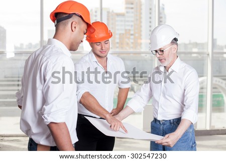 Cheerful workers are planning to build a construction. Senior architect is holding a blueprint. The foreman is pointing his finger at it. The men are looking at the paper and smiling