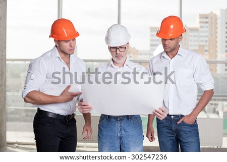 Grey-headed architect is holding a blueprint and looking at it seriously. He is explaining to his young colleagues the concepts of building. The workers are listening to his attentively