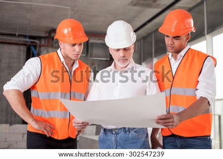 Successful old architect and two workers are discussing the plan of building. They are looking at the blueprint with seriousness