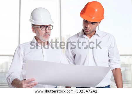 Experienced old architect and handsome young foreman are discussing the project. They are looking at the blueprint seriously. The men are wearing helmets