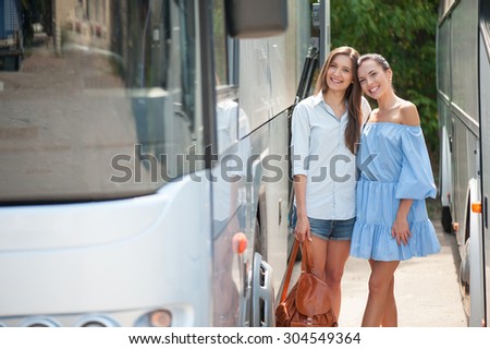 Cheerful women are standing between two buses and waiting for its departure. They are ready for traveling. The girls are embracing and smiling