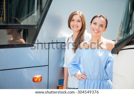 Attractive women are enjoying their trip. They are standing near a public transport. The friend are looking at the camera with joy and smiling