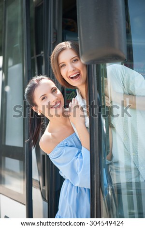 Pretty girls are standing on doorsteps of a bus. They are embracing and laughing. The girls are looking through the doors of vehicle with joy